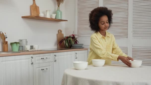 Malá Afroameričanka odstraní ze stolu hluboké bílé misky a umístí je do dřezu, aby se umyly. Konec jídla. Dospívající dívka pózuje na pozadí světlé kuchyně. Zpomalený pohyb. — Stock video