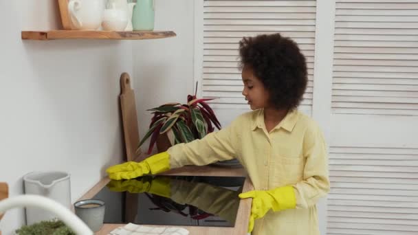 Kis afro-amerikai lány visel sárga gumikesztyűt mosóronggyal törli felülete elektromos tűzhely.Tini lány pózol a háttérben a világos konyha. Lassú mozgás.. — Stock videók
