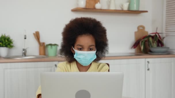 Klein Afrikaans-Amerikaans meisje met medisch masker gebruikt laptop voor online training of communicatie in sociale netwerken. Tienermeisje zit aan tafel tegen de achtergrond van een heldere keuken. Langzame beweging. — Stockvideo