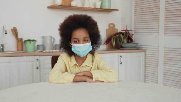 Kleines afroamerikanisches Mädchen mit medizinischer Maske blickt in die Kamera. Quarantäne, soziale Isolation im 19. Lebensjahr. Teenager Mädchen sitzt am Tisch vor dem Hintergrund der hellen Küche. Zeitlupe. — Stockvideo