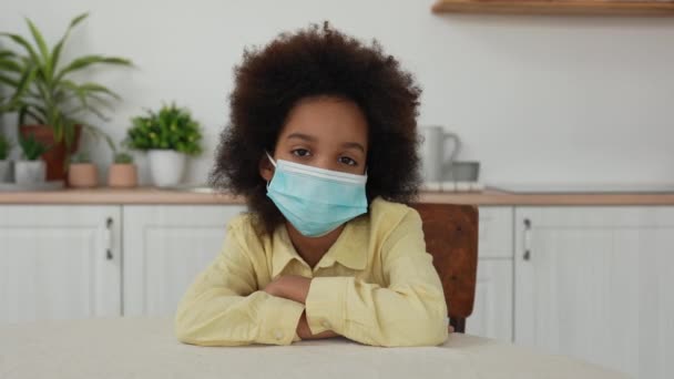 En liten afroamerikansk flicka i medicinsk mask tittar in bakom kameran. Tonårstjej sitter vid bordet mot bakgrund av ljust kök med vacker interiör. Sakta i backarna. Närbild. — Stockvideo