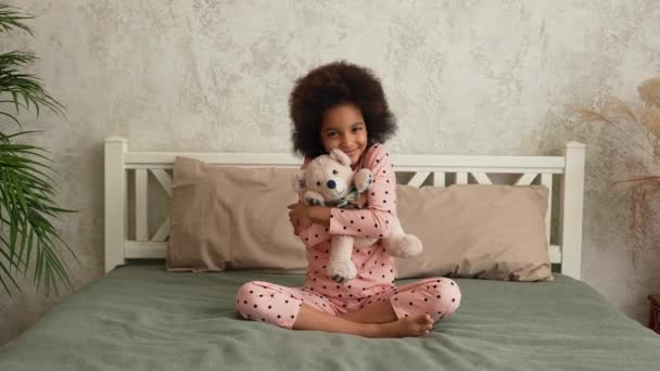 Schattig Afrikaans Amerikaans meisje knuffelt haar favoriete teddybeer. Tienermeisje in pyjama zit op bed in slaapkamer met prachtig licht interieur. Langzame beweging. — Stockvideo