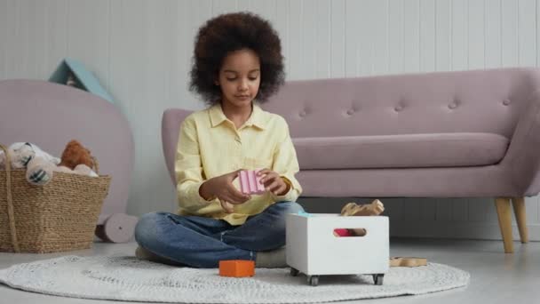 Malá Afroameričanka staví věž z barevné dřevěné stavebnice. Teenage dívka se hraje s hračkami, zatímco sedí na podlaze v dětském pokoji s krásným světlým interiérem. Zpomalený pohyb. — Stock video