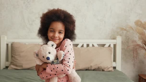 Petite fille afro-américaine mignonne étreint son ours en peluche préféré. Adolescente en pyjama se trouve sur le lit dans la chambre avec un bel intérieur clair. Au ralenti. Gros plan. — Video