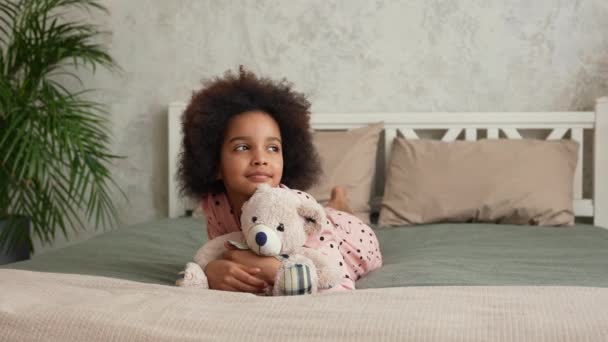Linda niña afroamericana abraza a su oso de peluche favorito. Chica adolescente en pijama se encuentra en la cama en el dormitorio con hermoso interior de luz. Movimiento lento. — Vídeo de stock