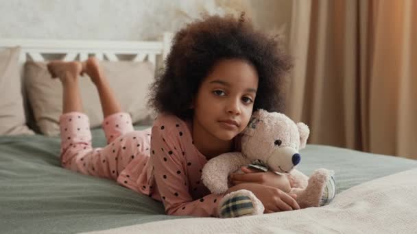 可爱的非洲裔美国小女孩抱着她最喜欢的泰迪熊。穿着睡衣的少女躺在床上，卧室里光线清澈，很美。慢动作靠近点. — 图库视频影像