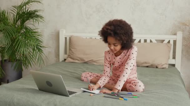 Söt liten afroamerikansk flicka upptagen med att skriva eller lära sig att rita med filt tips pennor genom att titta på bärbar dator. Tonårstjej i pyjamas sitter på sängen i sovrum med vacker ljus interiör. Långsamma rörelser — Stockvideo
