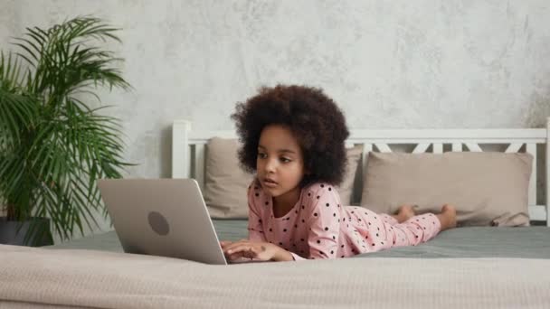 Cute little African American girl uses laptop to view information, communicate with friends in chat or social networks. Teenage girl in pajamas lies on bed in bedroom. Slow motion. — Stock Video