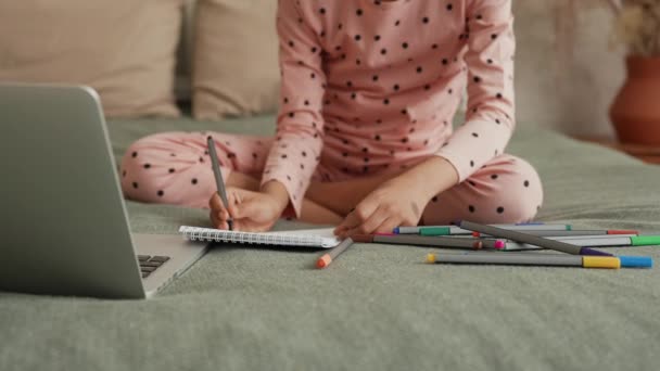 Cute little African American girl uczy się rysować z felttip długopisy patrząc na laptopa. Nastoletnia dziewczyna w piżamie siedzi na łóżku w sypialni z pięknym wnętrzem światła. Zwolnij trochę. Zamknij się.. — Wideo stockowe