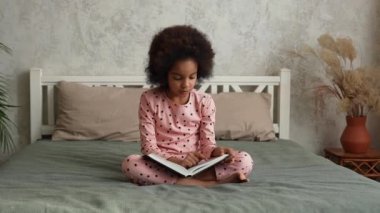 Yüzünde şaşkın ve coşkulu bir ifade olan peri masallarıyla kitap okuyan tatlı Afrikalı küçük bir kız. Pijamalı genç kız yatak odasında yatakta oturuyor. Yavaş çekim.