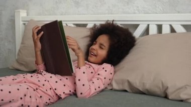 Şirin küçük Afrikalı Amerikalı kız peri masallarıyla dolu bir kitap okuyor ve uykuya dalıyor. Pijamalı genç bir kız yatak odasında uzanmış, içi aydınlık. Ağır çekim. Kapat..