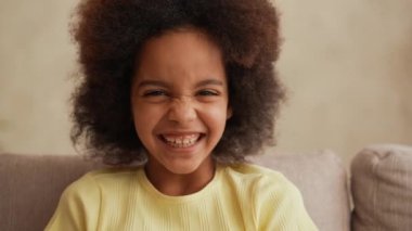 Küçük Afrikalı Amerikalı bir kızın portresi kameraya bakıp neşeyle gülüyor. Genç kız koridordaki gri koltukta oturuyor. Parlak, güzel bir odanın arka planına karşı. Ağır çekim. Kapat..