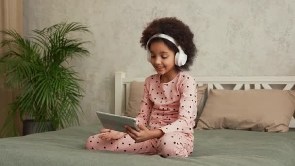 Schattig Afrikaans Amerikaans meisje in grote witte koptelefoons die praten op een video gesprek met behulp van een digitale tablet. Tienermeisje in pyjama zit op bed in slaapkamer met prachtig licht interieur. Langzame beweging. — Stockvideo