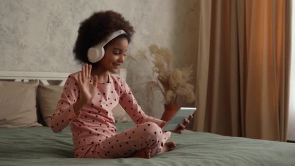 Schattig Afrikaans Amerikaans meisje in grote witte koptelefoons die praten op een video gesprek met behulp van een digitale tablet. Tienermeisje in pyjama zit op bed in slaapkamer met prachtig licht interieur. Langzame beweging. — Stockvideo