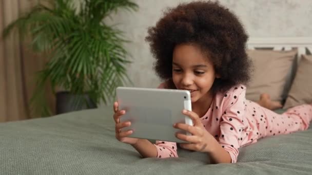 Menina afro-americana bonito em joga um jogo em um tablet digital e se alegra com a vitória. Menina adolescente em pijama encontra-se na cama no quarto com belo interior de luz. Movimento lento. — Vídeo de Stock