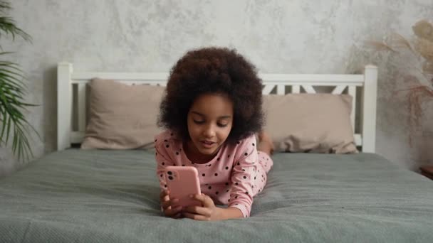 かわいいアフリカ系アメリカ人の女の子がスマートフォンの画面を見て歌い、ビデオを記録します。パジャマ姿の10代の少女は、美しい光のインテリアのベッドルームのベッドルームに横たわっています。スローモーション. — ストック動画