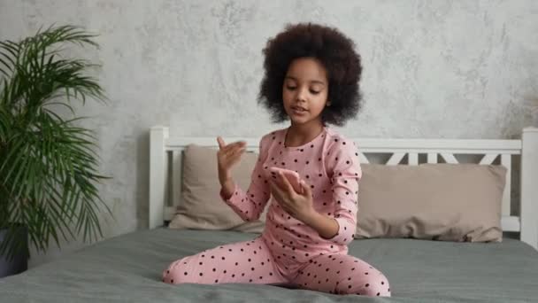 Mignonne petite afro-américaine chante en regardant l'écran du smartphone et enregistre la vidéo. Adolescente en pyjama se trouve sur le lit dans la chambre avec un bel intérieur clair. Mouvement lent. — Video