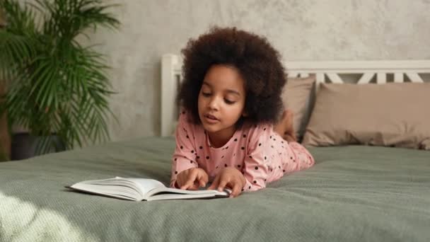 Leuk Afrikaans Amerikaans meisje leest enthousiast een boek met sprookjes. Tienermeisje in pyjama ligt op bed in slaapkamer met prachtig licht interieur. Langzame beweging. — Stockvideo