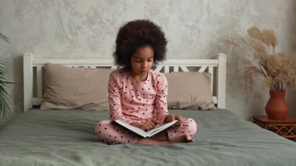 Linda niña afroamericana leyendo libro con cuentos de hadas con wow expresión sorprendida y entusiasta en su cara. Adolescente en pijama se sienta en la cama en el dormitorio. Movimiento lento. — Vídeo de stock