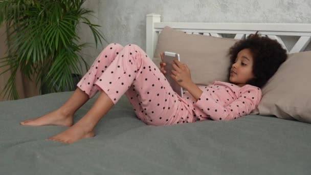 Cute little African American girl in plays a game on a digital tablet. Teenage girl in pajamas lies on bed in bedroom with beautiful light interior. Slow motion. — Stock Video