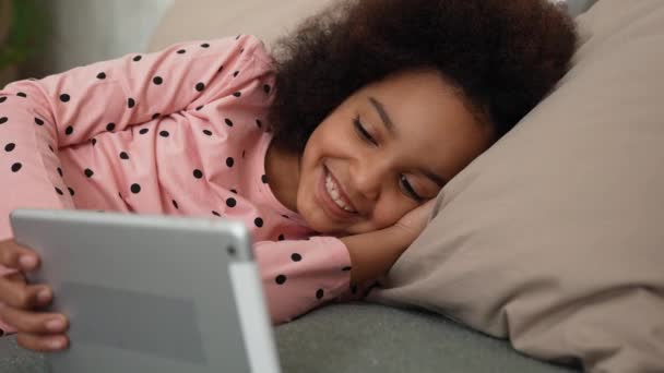 Schattig Afrikaans Amerikaans meisje dat met een digitale tablet naar een tekenfilm of sprookje kijkt. Tienermeisje in pyjama ligt op bed in slaapkamer met prachtig licht interieur. Langzame beweging. Sluiten.. — Stockvideo