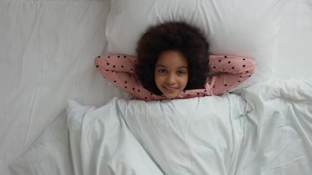 Bovenaanzicht van klein schattig Afrikaans Amerikaans meisje liggend onder deken en kijkend naar camera met glimlach. Tienermeisje in pyjama ligt op wit bed in slaapkamer met licht interieur. Langzame beweging. Sluiten.. — Stockvideo