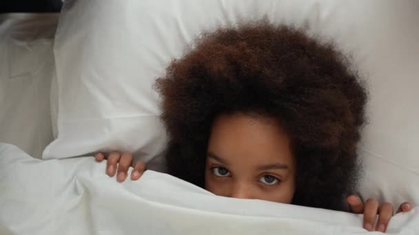 Şirin küçük Afrikalı Amerikalı kız yorganın altına saklanıyor, kafasıyla kapanıyor ve sonra ortaya çıkıyor. Pijamalı genç kız yatak odasında beyaz yatakta yatıyor. Ağır çekim. Kapat.. — Stok video