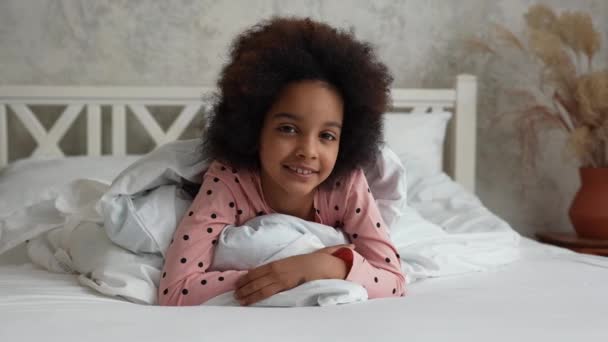 Retrato niña afroamericana tumbada bajo las sábanas, mirando a la cámara y sonriendo. Chica adolescente en pijama se encuentra en la cama blanca en el dormitorio con hermoso interior de luz. En cámara lenta. De cerca.. — Vídeos de Stock