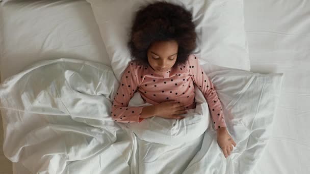 Top view of a little sleeping African American girl waking up stretching and yawning. Teenage girl in pajamas lies on white bed in bedroom. Slow motion. — Stock Video
