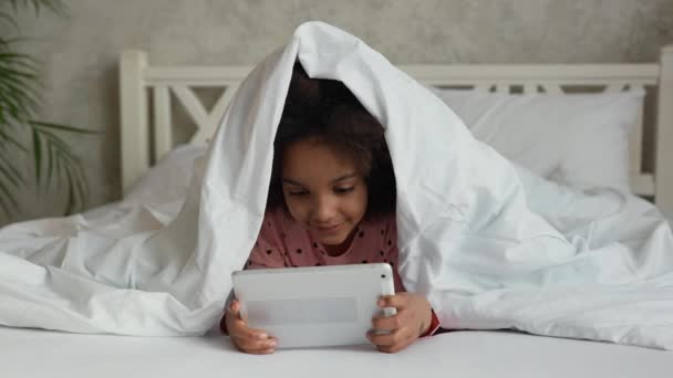 Portrait petite fille afro-américaine regarde dessin animé sur tablette numérique, a peur et se cache sous la couverture avec sa tête. Adolescente en pyjama se trouve sur lit blanc dans la chambre. Au ralenti. Gros plan. — Video