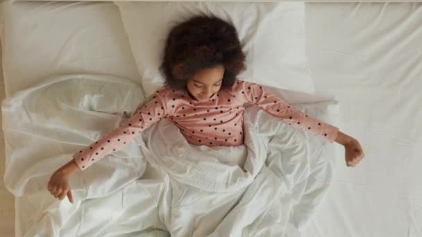 Vista dall'alto di una piccola ragazza afroamericana addormentata che si sveglia allungando e sbadigliando. Adolescente in pigiama si trova sul letto bianco in camera da letto. Rallentatore. — Video Stock
