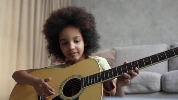 ノートパソコンの画面を見ながらギターを学ぶアフリカ系アメリカ人の少女。明るい美しい部屋のインテリアを背景に、 10代の女の子がホールの床に座っています。スローモーション。閉じろ!. — ストック動画