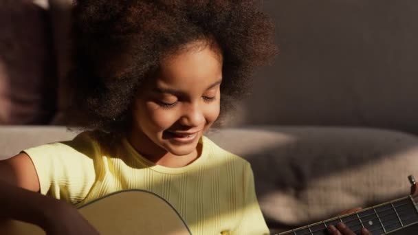 Retrato pequena menina afro-americana toca uma guitarra em raios de sol brilhante. Menina adolescente senta-se no chão no salão contra o pano de fundo de um belo interior quarto brilhante. Movimento lento. Fechar. — Vídeo de Stock