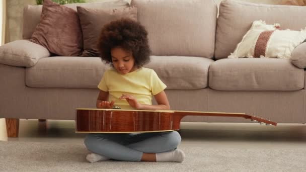 Porträtt lilla afroamerikanska flicka kranar handflator på ytan av trä gitarr skapa musikalisk rytm. Tonårstjej sitter på golvet i hallen mot bakgrund av ljusa vackra rum interiör. Långsamma rörelser — Stockvideo