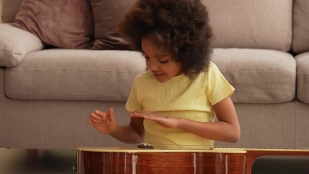 Portret kleine Afro-Amerikaanse meisje tikt handpalmen op het oppervlak van de gitaar creëren muzikale ritme. Tiener meisje zit op de vloer in de hal tegen de achtergrond van lichte kamer interieur. Langzame beweging. Sluiten.. — Stockvideo