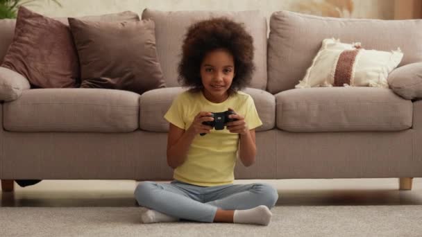 Portrait petite afro-américaine jouant à un jeu vidéo à l'aide d'une manette sans fil. Adolescente s'assoit sur le sol dans le hall dans le contexte d'une belle pièce intérieure lumineuse. Mouvement lent — Video