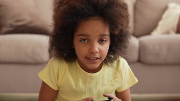 Portrait petite fille afro-américaine jouant à un jeu vidéo en utilisant une manette sans fil. Adolescente est assise sur le sol dans le hall dans le contexte d'une belle pièce intérieure lumineuse. Au ralenti. Gros plan. — Video