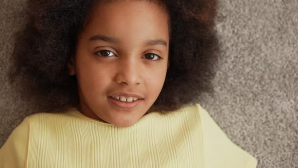 Die Kamera zoomt auf Gesicht und Augen eines kleinen afroamerikanischen Mädchens, das in die Kamera blickt und lächelt. Teenager-Mädchen liegt auf dem Boden vor dem Hintergrund eines grauen, flauschigen Teppichs. Zeitlupe. Nahaufnahme. — Stockvideo