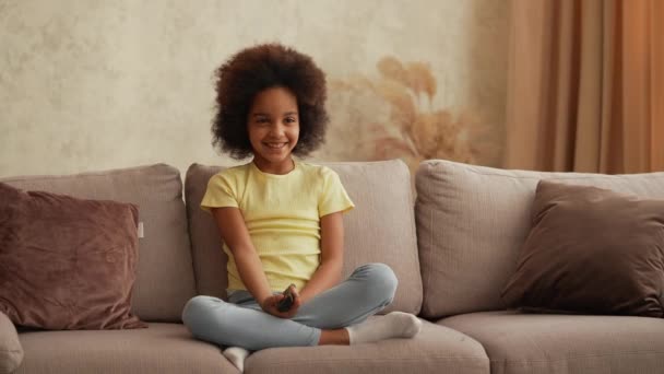 Portrait petite afro-américaine regarde la télévision et change de chaînes avec intérêt avec télécommande. Adolescente s'assoit sur un canapé gris dans le hall sur fond d'intérieur lumineux de la pièce. Mouvement lent. — Video