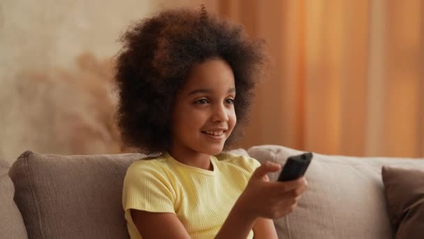 Portrait petite afro-américaine regarde la télévision et change de chaînes avec intérêt avec télécommande. Adolescente s'assoit sur un canapé gris dans le hall. Au ralenti. Gros plan. — Video