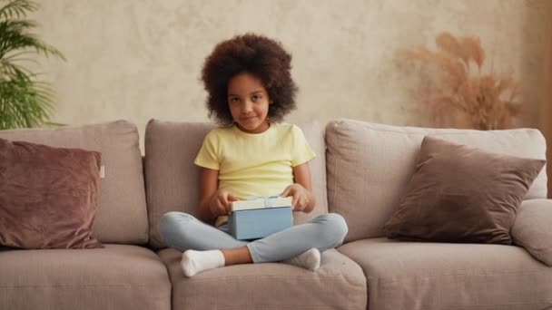 Portrait kleines afroamerikanisches Mädchen öffnet Geschenkbox, aufgebracht über das, was er sah, und öffnet dann Geschenk und ist sehr glücklich. Teenager-Mädchen sitzt auf grauem Sofa im Flur. Zeitlupe. — Stockvideo