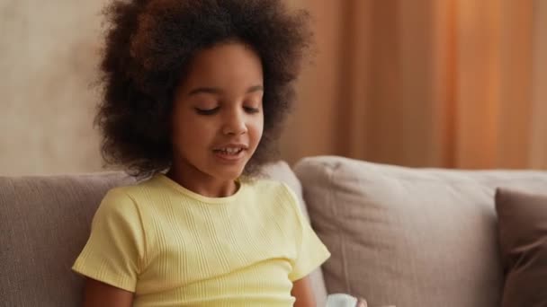 Portret klein Afro-Amerikaans meisje opent geschenkdoos en is erg blij. Tiener meisje zit op een grijze bank in de hal tegen de achtergrond van heldere mooie kamer interieur. Langzame beweging. Sluiten.. — Stockvideo