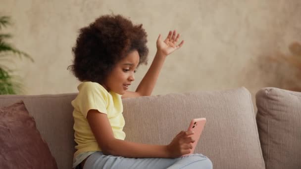 Portret kleine Afro-Amerikaanse meisje bekijkt informatie op een smartphone. Tiener meisje zit op een grijze bank in de hal tegen de achtergrond van heldere mooie kamer interieur. Langzame beweging. Sluiten.. — Stockvideo
