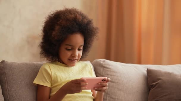 Portrait petite afro-américaine joue à un jeu sur smartphone. Adolescente s'assoit sur un canapé gris dans le hall contre toile de fond de lumineux bel intérieur de la chambre. Au ralenti. Gros plan. — Video