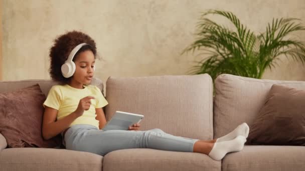 Porträtt lilla afroamerikanska flicka i stora vita hörlurar lyssnar musik med hjälp av digital tablett. Tonårstjej sitter på grå soffa i hallen mot bakgrund av ljusa vackra rum interiör. Långsamma rörelser — Stockvideo
