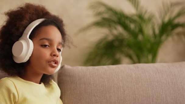 Портрет маленькой афроамериканской девочки в больших белых беспроводных наушниках слушает музыку. Девочка-подросток сидит на сером диване в холле на фоне яркого красивого интерьера комнаты. Медленное движение. — стоковое видео