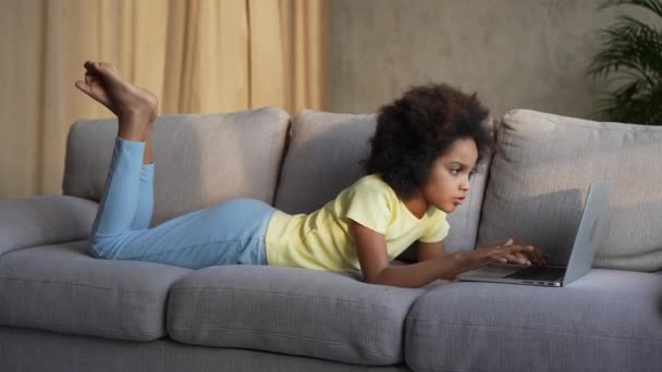 Vue de côté petite fille afro-américaine regarde l'information sur un ordinateur portable ou communique dans le chat en ligne, les réseaux sociaux. Adolescente se trouve sur un canapé gris dans le hall. Mouvement lent. — Video