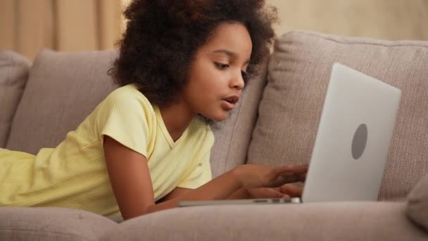 Vue de côté petite fille afro-américaine regarde l'information sur un ordinateur portable ou communique dans le chat en ligne, les réseaux sociaux. Adolescente se trouve sur un canapé gris dans le hall. Au ralenti. Gros plan. — Video