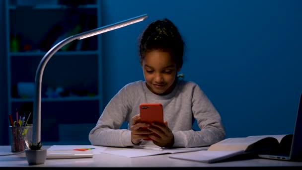 Een meisje gooit haar pen op tafel, vouwt haar armen over haar borst, pakt haar telefoon op, speelt spelletjes. Het kind zit aan tafel bij het licht van een nachtlampje. Sluit maar af. Slow motion klaar 59,97 fps. — Stockvideo