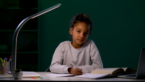 A menina olha atentamente para a câmera e libera a caneta de suas mãos. Uma rapariga a fazer trabalhos de casa sentada à mesa à luz de um candeeiro nocturno. Fecha. Movimento lento pronto 59.97fps. — Vídeo de Stock
